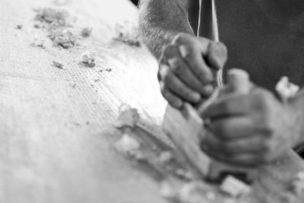 Close up of carpenter's hands using a hand plane for smoothing a plank of wood in his workshop. - black n white -