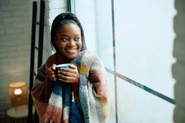 istock Happy black woman wrapped in a blanket drinking tea by the window. 2177133056