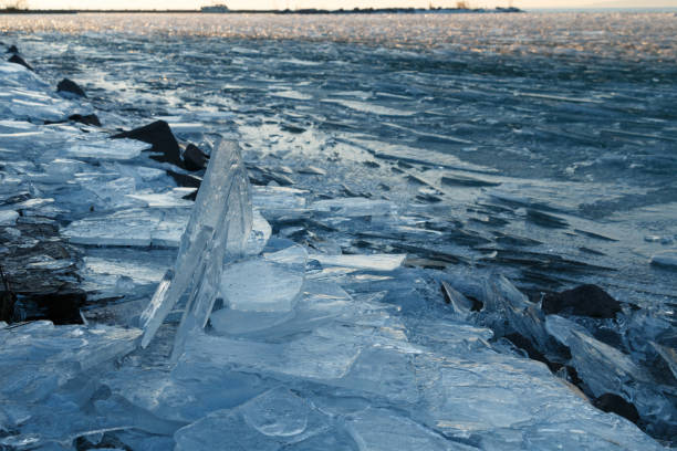 A frozen lake covered with cracked ice sheets near a rocky shore, with the icy surface stretching towards the horizon. The scene is bathed in cool, natural light under a clear sky, with distant hills visible in the background.