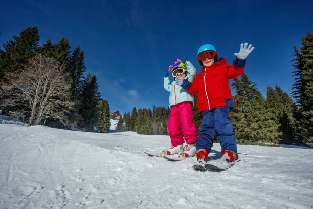 A pair of youngsters, equipped with ski and helmets, enjoy the snow-covered slope, a backdrop of tall trees a brilliant blue sky on background