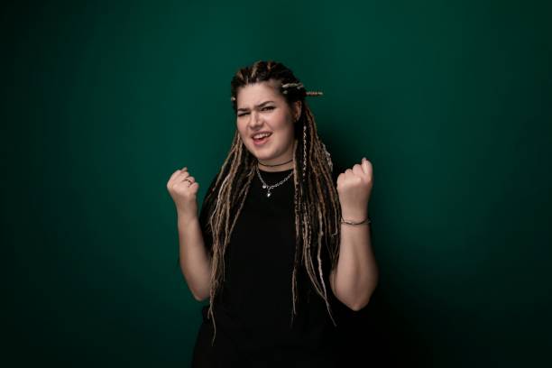 A woman with dreadlocks is standing against a vibrant green wall. She is looking confidently at the camera, showcasing her unique hairstyle.