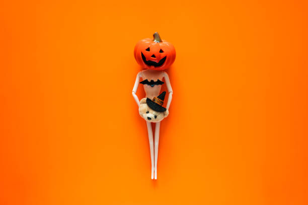 Halloween background with doll wearing Jack o lantern on head and holding small decoration on orange background. Minimal concept.