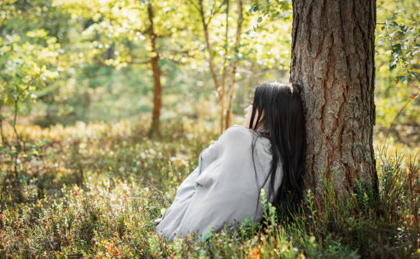 A woman in a light gray garment leans against the trunk of a tree, enjoying a moment of peace amidst vibrant greenery on a sunny day in the forest.