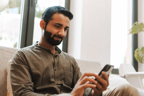 istock Smiling bearded Indian man holding mobile phone using mobile app, sitting on comfortable sofa 2177197147