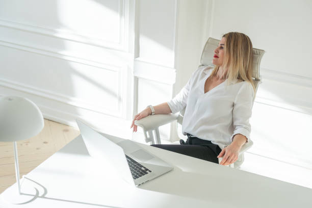 istock Delighted young blonde woman sitting at white table in blouse using laptop 2177200146