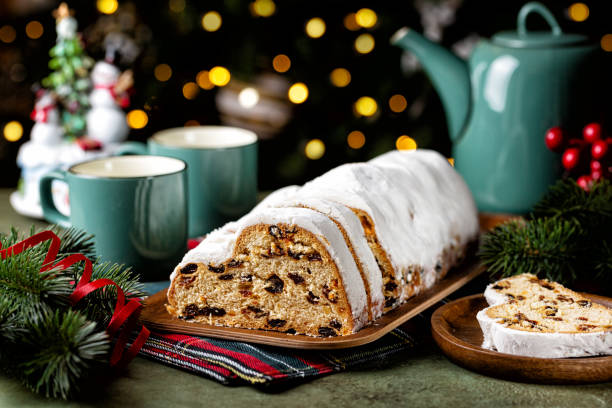 Served festive table with Stollen cake, tea cups and pot. German Christmas dessert, bread chock full of dried fruit, candied peel, and almonds. Festive lights, tree brunch and decoration.