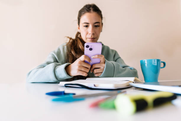 istock Female student using a cell phone while sitting at a table in the living room. 2177204172