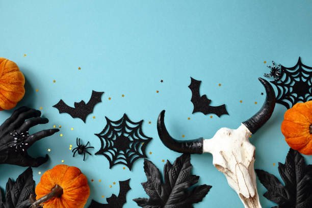 Halloween flat lay composition with scary decor, cow skull, pumpkins, cobwebs, spiders, bats, black maple leaves on blue background.