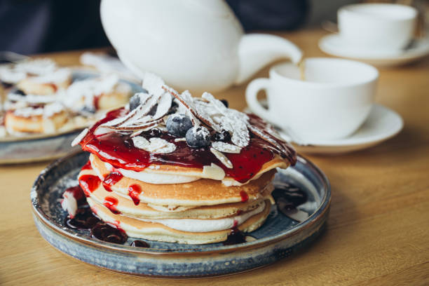 A tall stack of pancakes topped with coconut slices and blueberries, drizzled with thick jam. Breakfast pancakes with berries in cafe. Enjoy a delightful morning with a generous stack of pancakes.