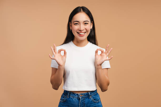 istock Asian woman smiling and gesturing ok sign with both hands looking at camera on beige background 2177231701