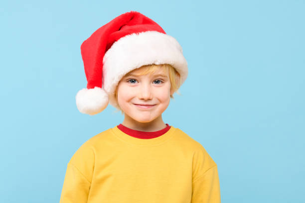 Cute young caucasian preschooler boy wearing santa hat, smiling while looking at camera. Studio portrait isolated over pastel blue background.