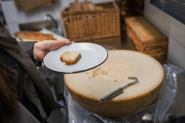 A person stands beside a large cheese wheel, holding a plate with a slice of bread, suggesting a culinary experience or tasting session. The setting appears cozy and inviting.