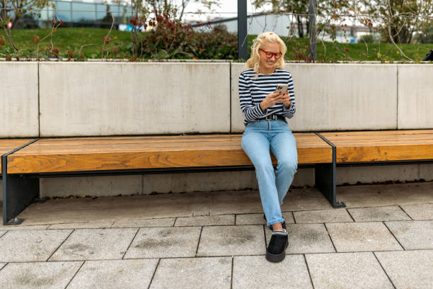 istock Smiling woman sitting on bench with smartphone in hand 2177261889