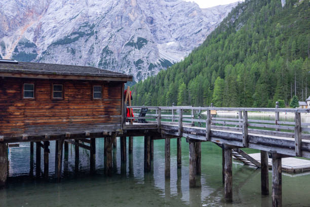 A rustic wooden cabin on stilts extends over the calm waters of a mountain lake, connected by a wooden pier. The scene features lush forests and towering rocky peaks in the background.