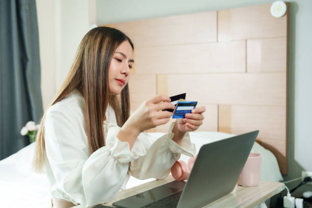 Asian woman comparing multiple credit cards while online shopping using laptop sitting in bedroom wearing white blouse modern e-commerce lifestyle focused consumer financial concept. mujer asiática que compara varias tarjetas de crédito mientras compra en línea usando una computadora portátil sentada en el dormitorio con blusa blanca estilo de vida moderno centrado en el comercio electrónico, concepto financiero del consumidor.