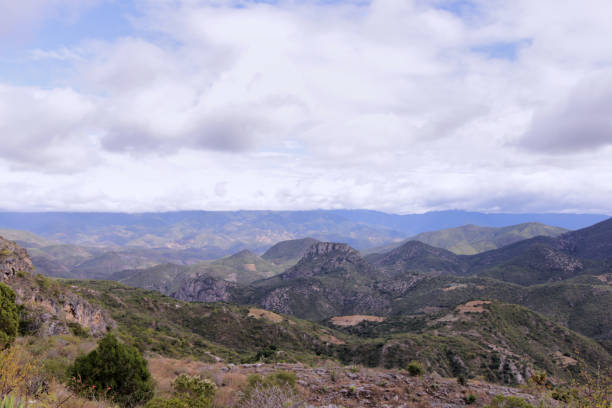 A breathtaking view of the rolling hills and mountains in Oaxaca, Mexico, under a cloudy sky. The landscape features varying shades of green, with lush vegetation and rocky formations interspersed throughout the scene. In the distance, the mountains create a dramatic backdrop, and patches of open land reveal hints of agricultural fields. The clouds overhead add depth to the sky, suggesting a change in weather and enhancing the natural beauty of this serene mountainous region. This image encapsulates the tranquility and rugged charm of Oaxaca’s highlands.