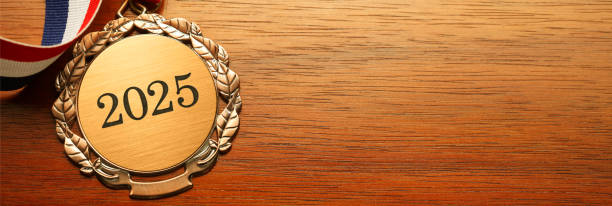 A close up of a gold medal engraved with the year 2025 and an attached red, white, and blue ribbon resting on a wood table.