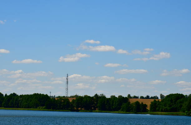A view of a vast river or lake surrounded from all sides with reeds, forests, moors, fields, and pasturelands located next to a small marina or wooden platform and some metal tower seen in Poland