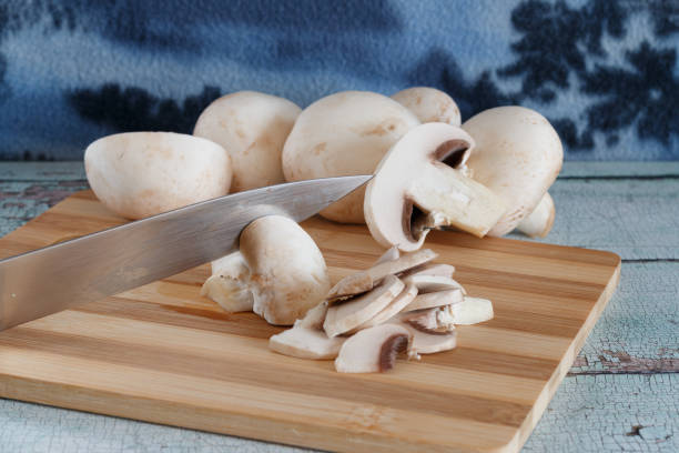A close-up view fresh white mushrooms on a wooden cutting board. Several whole mushrooms and tomatoes are visible in the background, highlighting the preparation of fresh ingredients