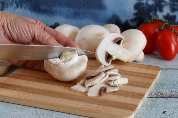 A close-up view of a hand slicing fresh white mushrooms on a wooden cutting board. Several whole mushrooms and tomatoes are visible in the background, highlighting the preparation of fresh ingredients.