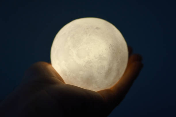 istock Hands holding an illuminated moon-like sphere lamp against a dark backdrop 2177318143