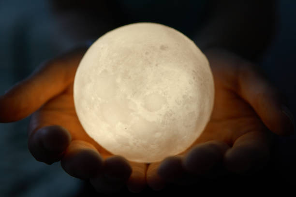 istock Hands holding an illuminated moon-like sphere lamp against a dark backdrop 2177318230