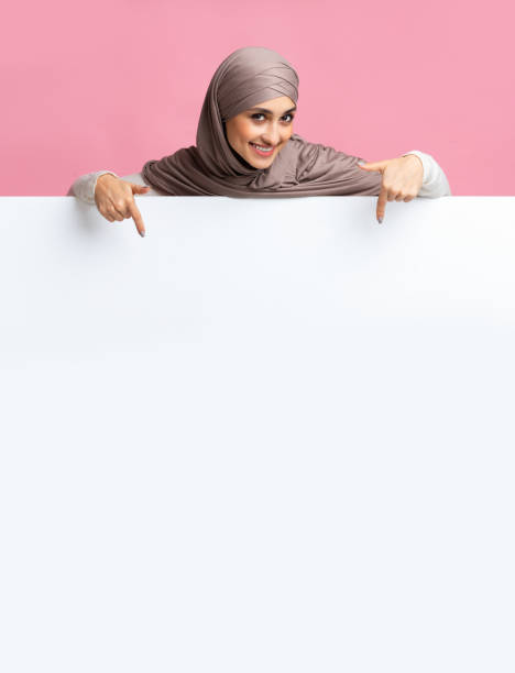 istock Cheerful arabic girl in headscarf pointing at white blank advertisement board 2177321766