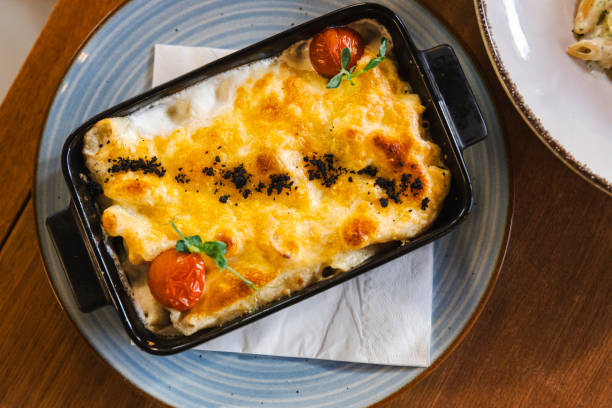 Deliciously baked macaroni topped with melted cheese and garnished with fresh tomatoes. Served in a black dish on a blue plate, it's a tempting and savory meal option.