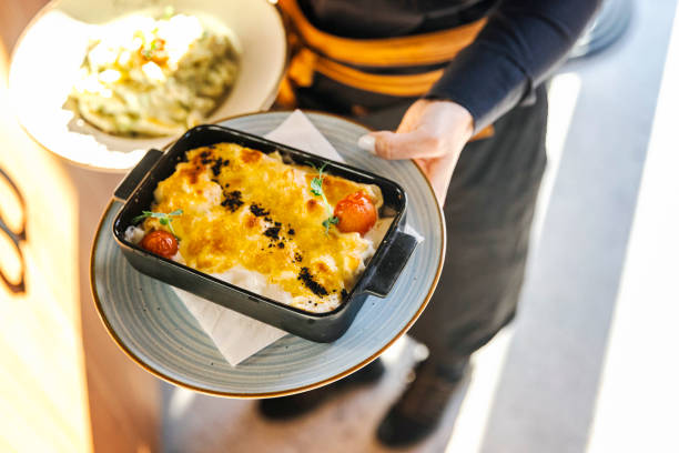 Close-up of a savory cheese casserole garnished with cherry tomatoes, held by a server in a cozy restaurant ambiance, highlighting the art of culinary presentation.