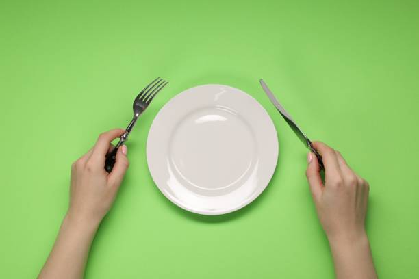 istock Woman holding fork and knife near empty plate at green table, top view 2177326488
