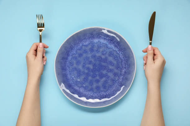 istock Woman holding fork and knife near empty plate at light blue table, top view 2177326503
