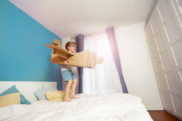 istock Child imagines flying with a cardboard airplane, standing on bed 2177333622