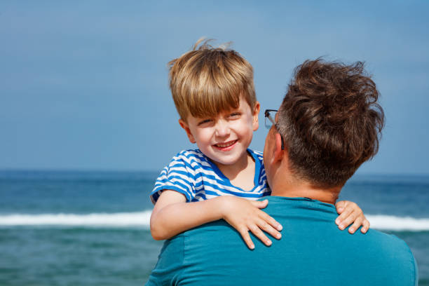 A cheerful child in a striped shirt is carried by an adult parent near the sea, as waves roll in behind them, happy vacation moment