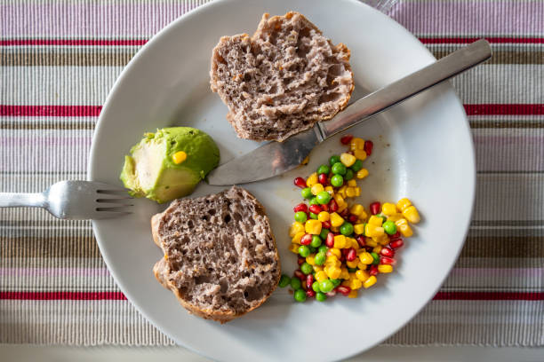 A white dinner plate with a bread roll, and avocado half, and peas and corn and pomegranate salad.