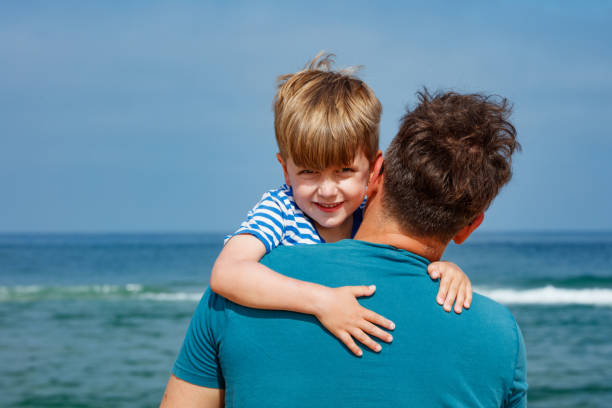 On a sunny day at the beach, a happy boy grins while his father hold him, and the ocean is visible in the background