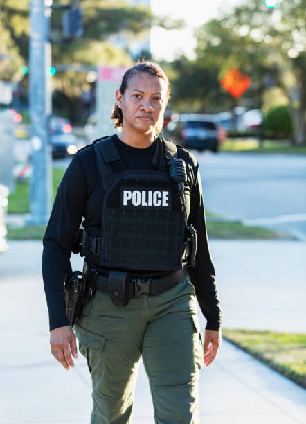 A mature multiracial policewoman patrolling an urban area on foot. She has a serious expression as she walks on a sidewalk, looking toward the camera.