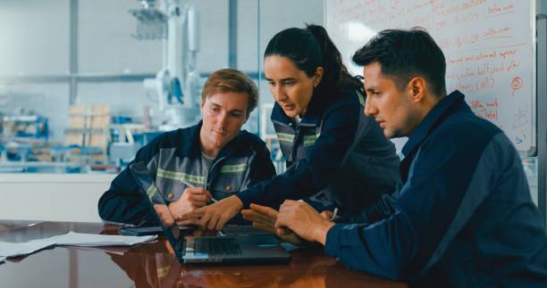 Group of engineers in factory meeting, collaborating and analyzing data on laptop, focusing on project development and teamwork. Robotic Industrial Factory.