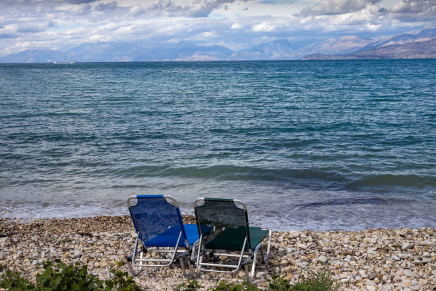 Two blue empty deck chairs on a pebble beach of the Ionic sea. Mountains in Albania on the horizon. Intense clouds in the late summer. North-East Corfu, Greece.