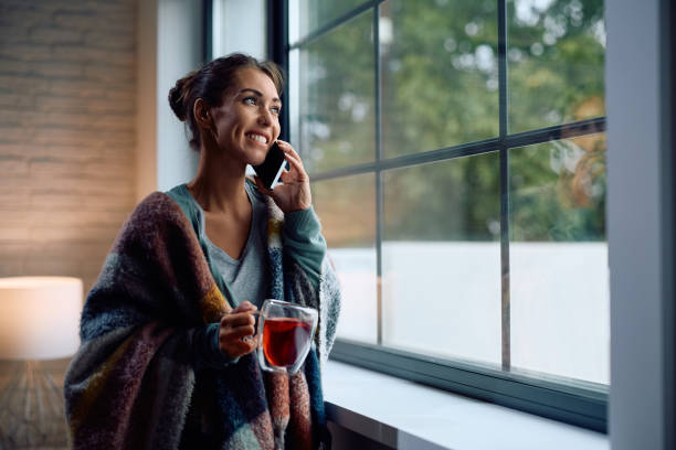 istock Smiling woman wrapped in a blanket drinking tea while talking on the phone by the window. 2177401388
