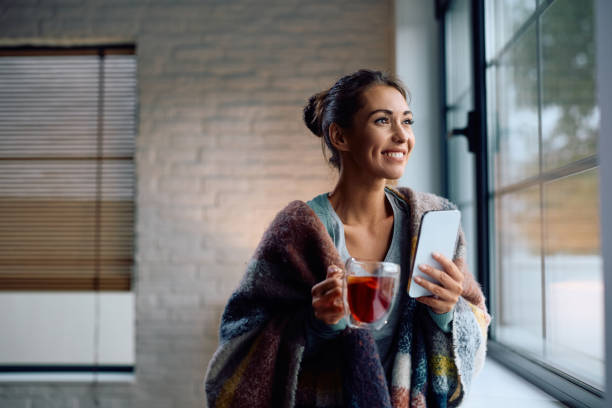 istock Happy woman drinking tea while using smart phone by the window at home. 2177401676