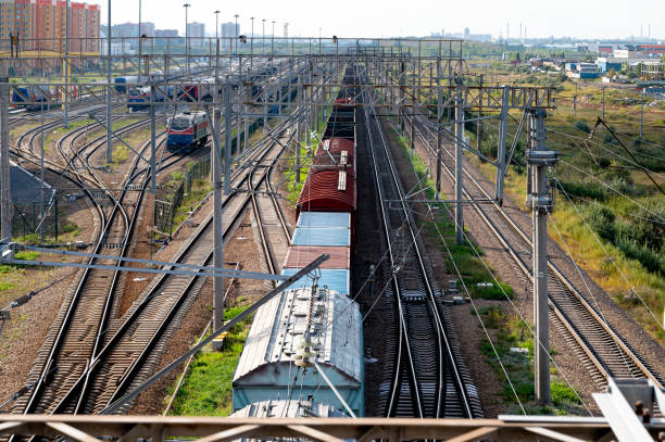 Freight trains on a busy railway junction with multiple tracks, switches, and overhead power lines, leading towards a distant city skyline and industrial area, representing transportation, logistics