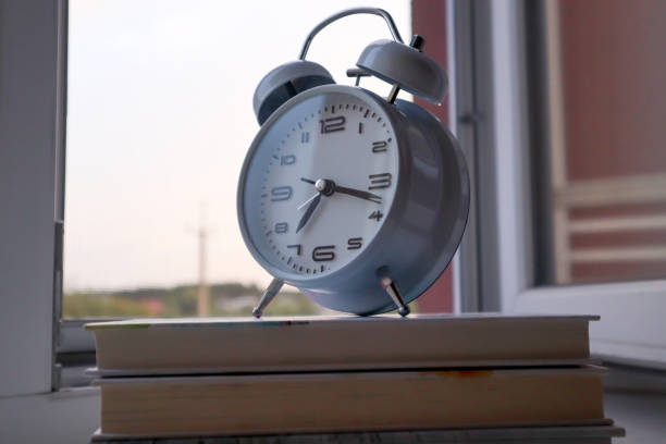 White retro alarm clock staying on a stack of books on the window sill. Time for reading.