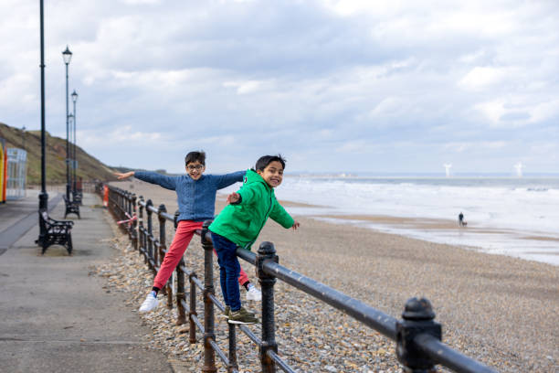 Wide shot of two young boys balancing on a promenade railing beside a pebble beach in Saltburn-by-the-Sea, North East England. They are smiling with their arms outstretched, wearing warm clothing on a winter's day. The sea is visible in the distance, waves breaking on the shore.\n\nVideos similar to this scenario available.