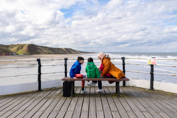 Wide shot of a mother and her two children sitting on a bench on a pier in Saltburn-by-the-Sea, North East England. They are smiling and talking, wearing warm clothing on a sunny winters day. In the background is an expanse of coastline with waves breaking on the shore.\n\nVideos similar to this scenario available.