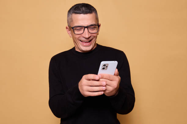 istock Cheerful guy wearing glasses and jumper smiling while browsing smartphone checking social media feeling happy connected with people through technology standing indoor isolated over beige background 2177451809