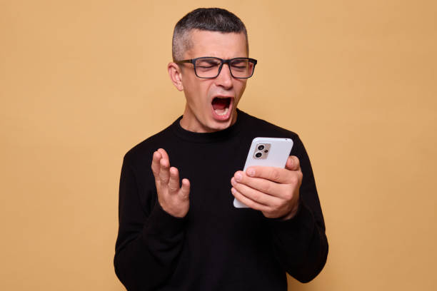 istock Angry man wearing glasses and jumper screaming while holding smartphone feeling aggressive about problems with internet connection exclaiming frustrations isolated over beige background 2177451814