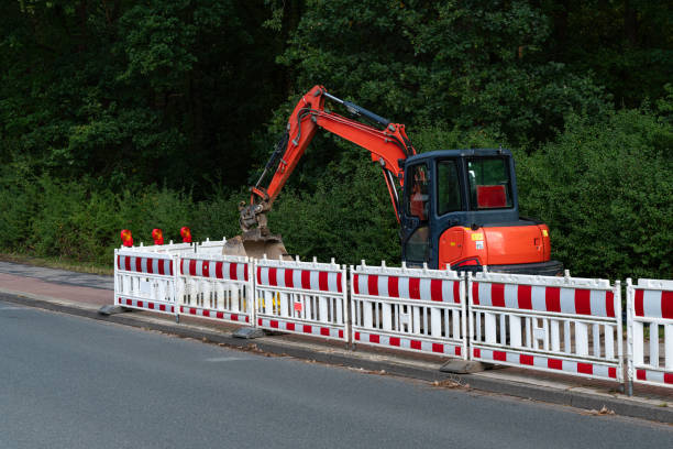 A construction crew is using an excavator to dig along the roadside near a wooded area during the evening. Safety barriers are set up to protect the site and passersby.