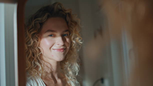 Smiling woman in mirror reflection in warm sunlight. Portrait happy blonde posing in home bathroom. Joyful relaxed girl looking admiring natural beauty indoors. Serene curly model enjoying leisure