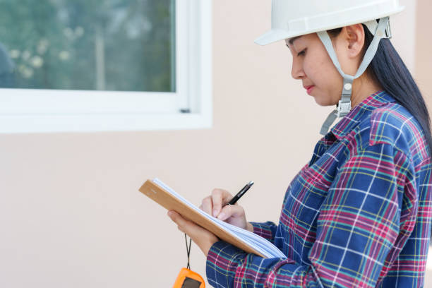 istock Asian female construction worker inspects new house windows, taking notes on clipboard, focusing on detail, wearing white helmet, standing outdoors on bright sunny day, final handover process. 2177462129