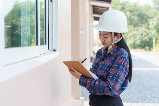 Asian woman wearing white safety helmet inspects window of new house before handover to customer holding clipboard and pen, checks construction details, outdoor setting, sunny day, working atmosphere. una mujer asiática con casco de seguridad blanco inspecciona la ventana de la nueva casa antes de entregarla al cliente sosteniendo un portapapeles y un bolígrafo, verifica los detalles de construcción, el entorno al aire libre, el día soleado, el ambi